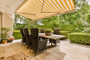 an outdoor dining area with table and chairs under a large yellow striped awning over the patio, surrounded by lush green trees